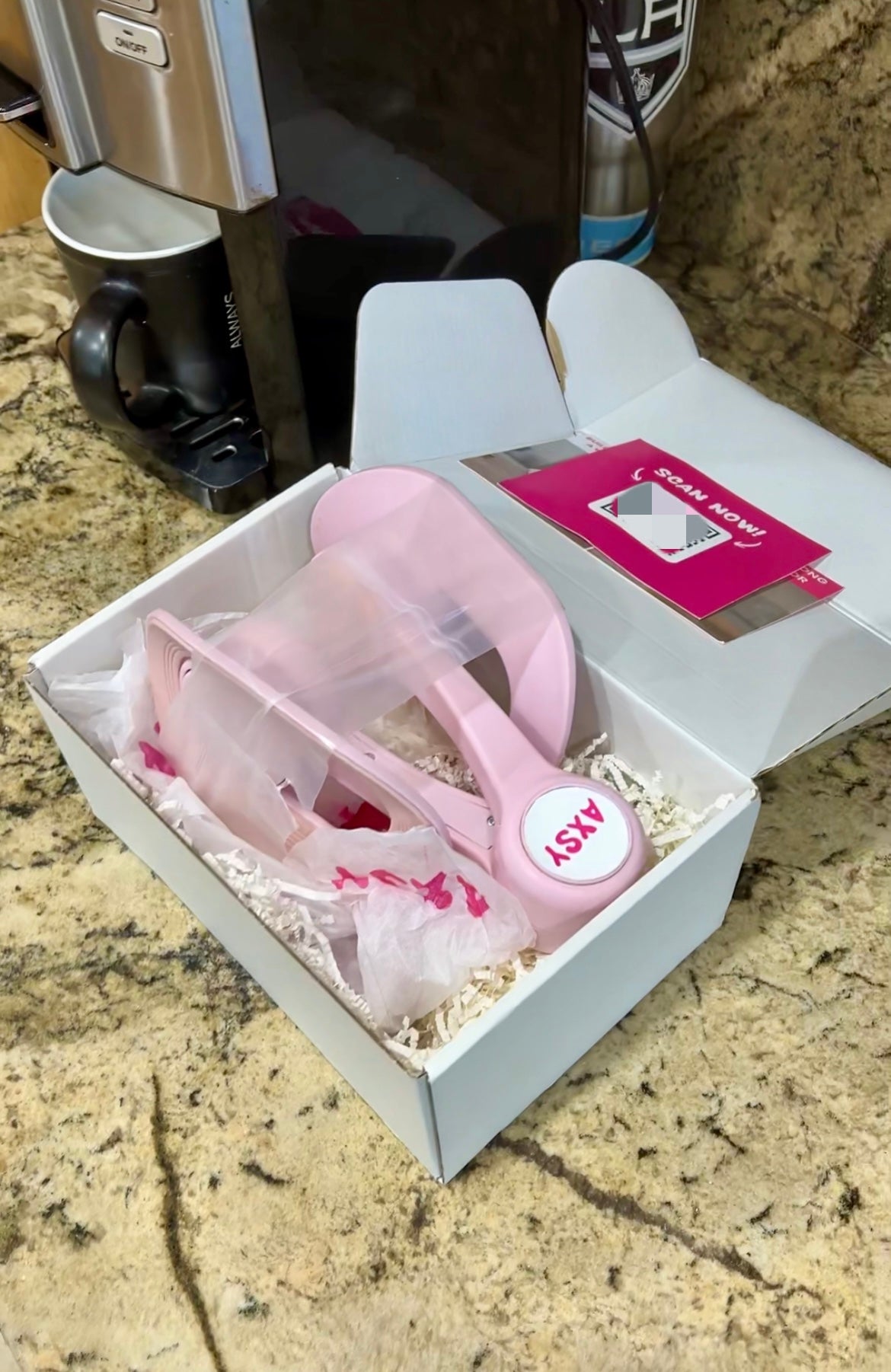 Open AXSY product box on a kitchen countertop with the pink pelvic floor and inner thigh trainer nestled in white crinkle paper. The package includes branded tissue paper and a QR code card, showcasing a premium unboxing experience with the trainer fully visible and ready to use